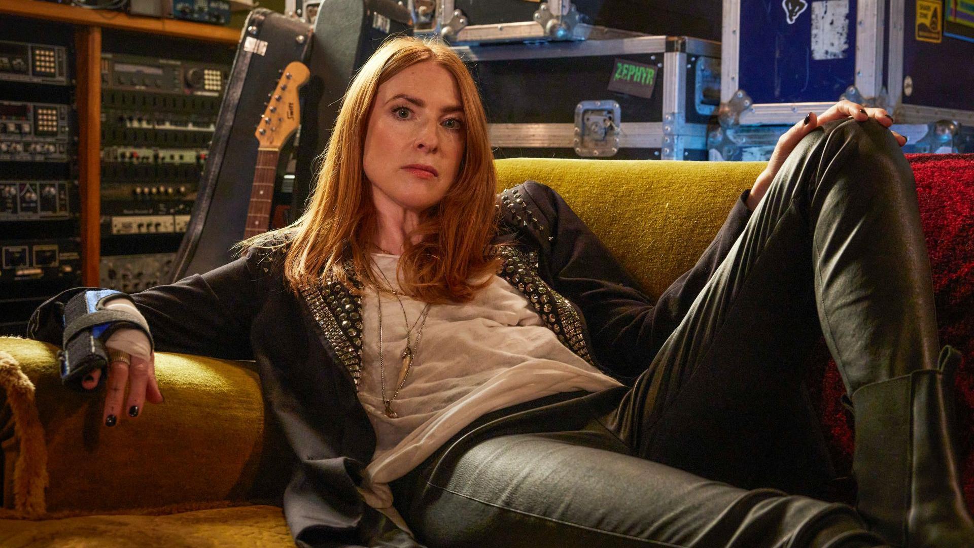 Rosalie Craig lies on a orangey brown sofa. She is wearing black leather trousers a black jacket and a white t-shirt. Behind her is a guitar propped against a collection of recording equipment and several instrument and amp cases.