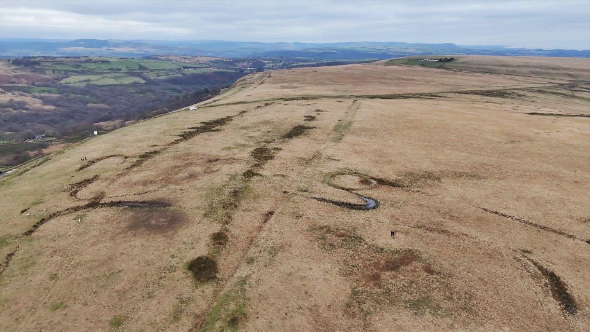 A photograph of the Tor Clawdd ring cairn shot from the air by a drone - the wider Mynydd y Gwair common is also visible - a large, flat expanse of brown grass and vegetation.