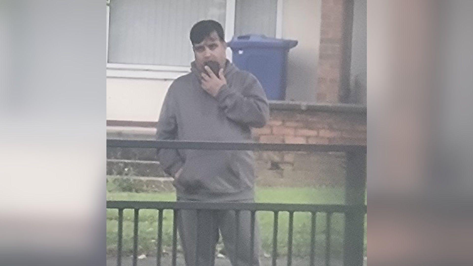 A man with mid-length dark hair wearing a grey tracksuit and standing at the side of a road. He has his phone to his mouth.