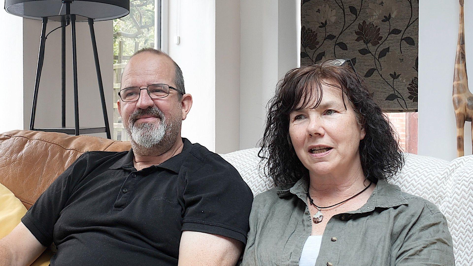 A man in a black polo shirt sits on a sofa next to a woman in a green shirt