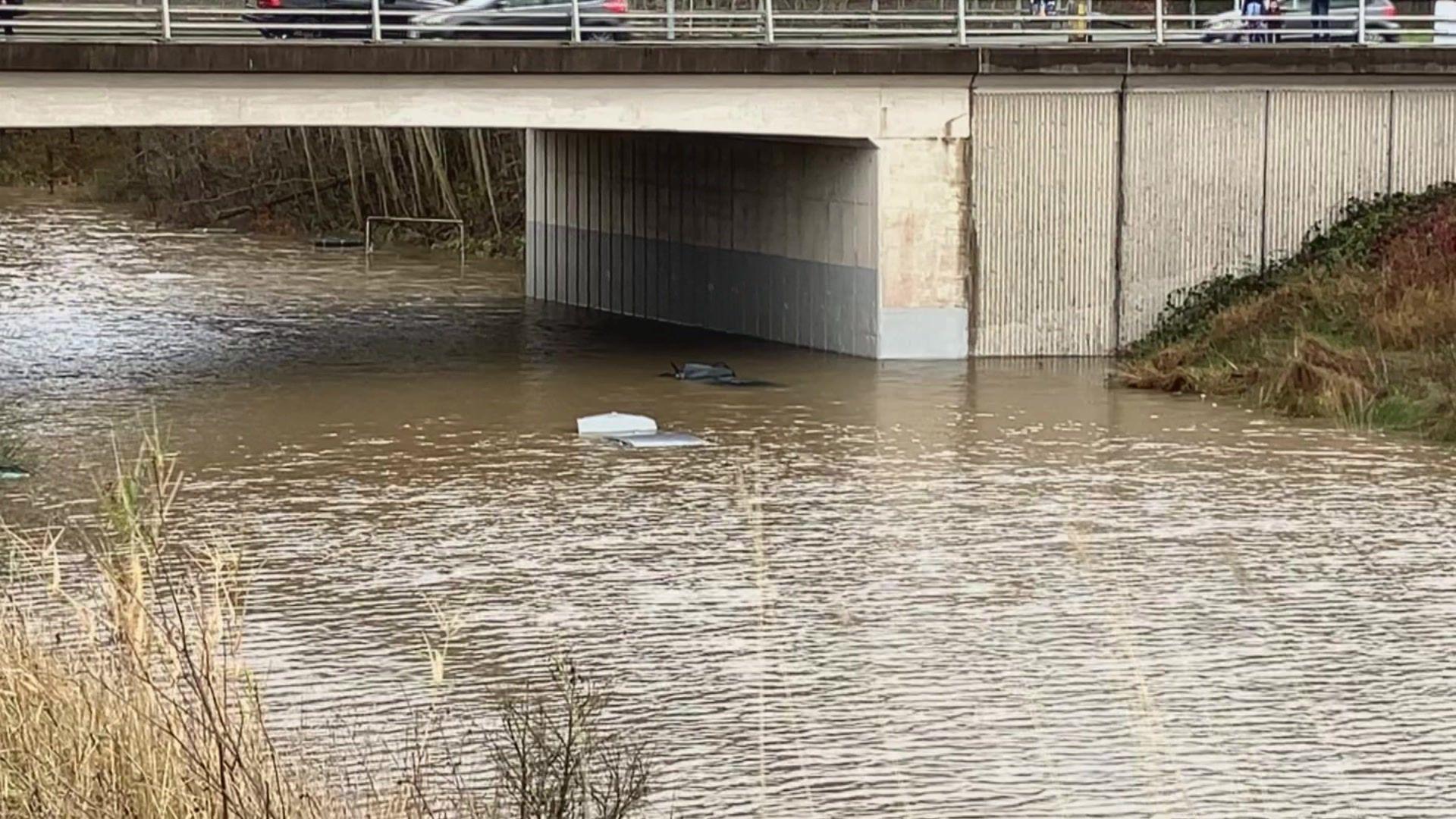 A road under a bridge is completely flooded, with the roofs of a grey and black car just about visible over the water's surface