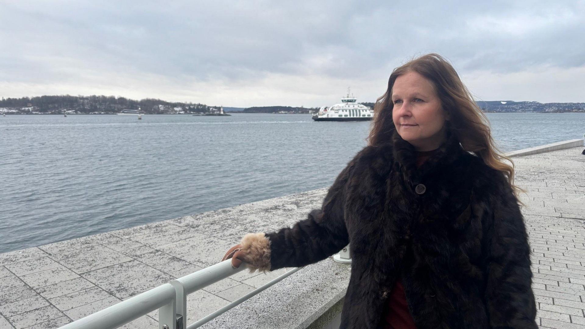 A woman in a brown coat stands looking over water on a cold, grey day in Oslo