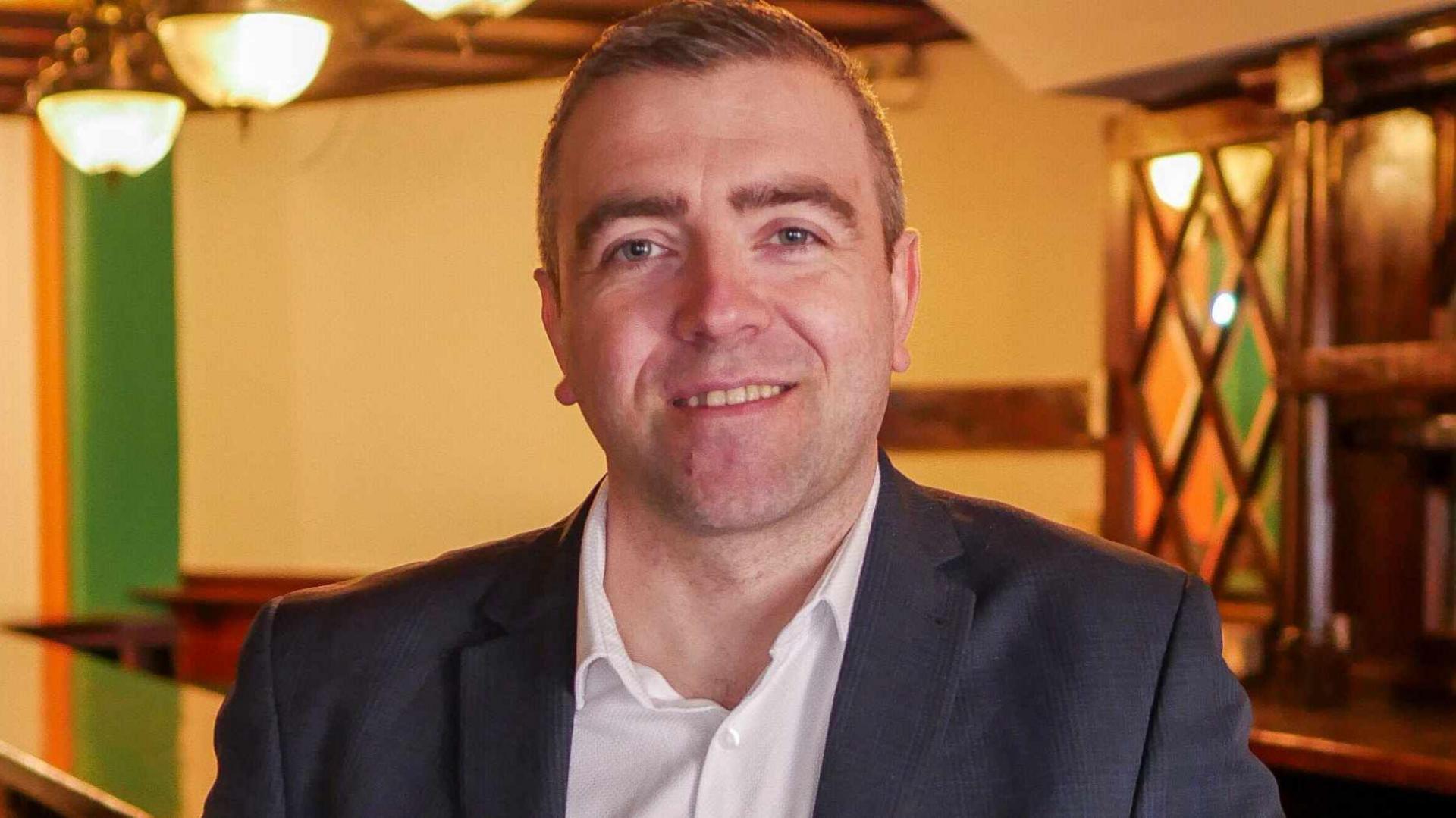 A man with short brown hair, a white shirt and a black formal jacket is stood indoors. He is smiling at the camera