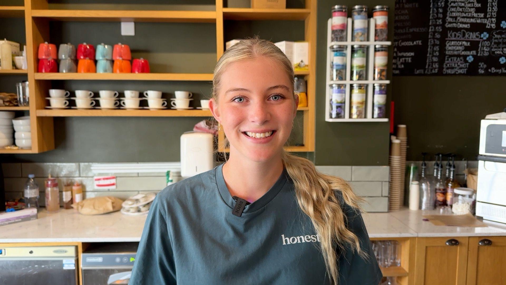 A young woman with long blonde hair, tied in a ponytail, and a bluey grey T-shirt, bearing the word "honesty" smiling and standing in from of a cafe's counter