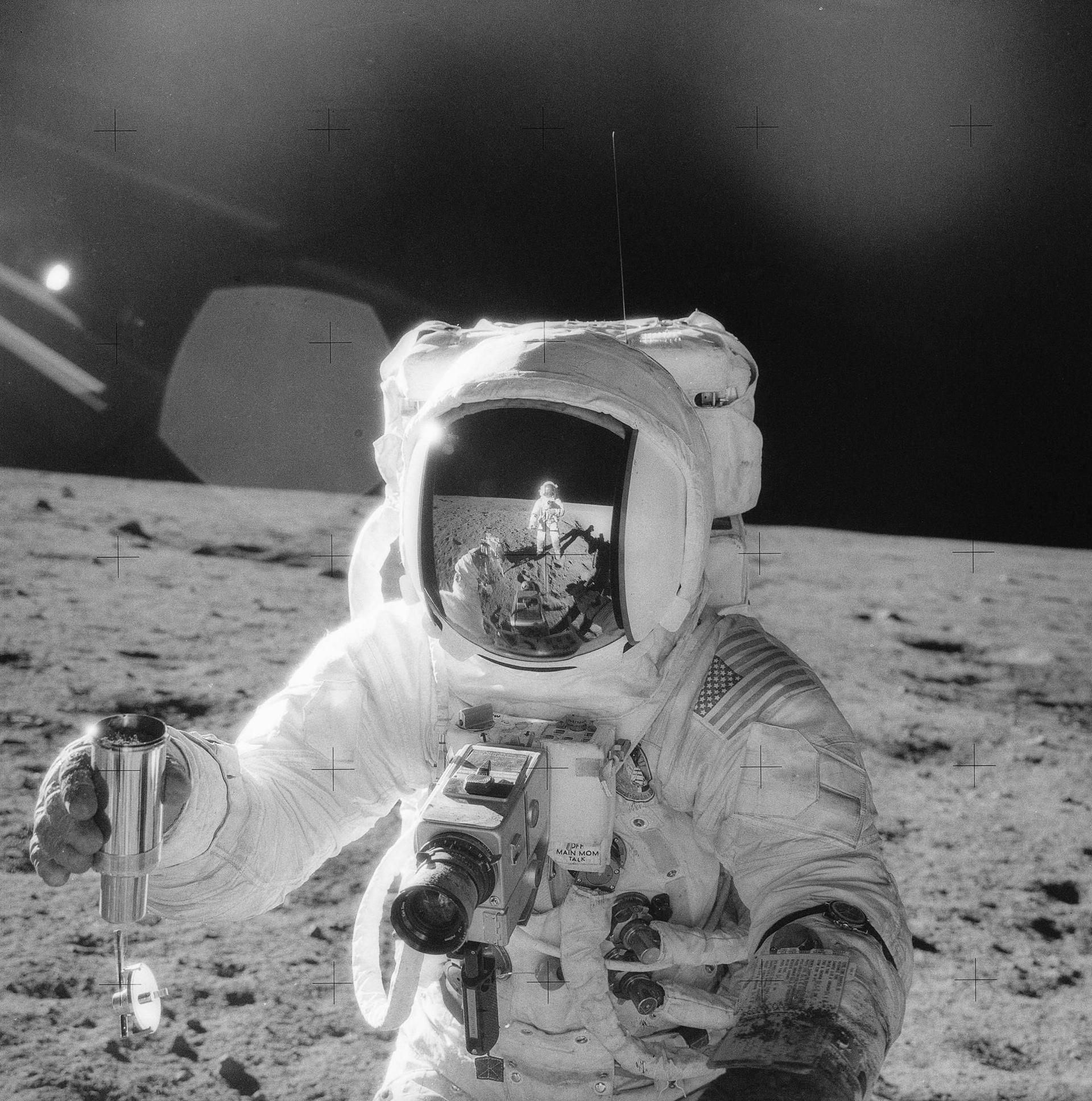 Black-and-white still of Apollo 12 mission in 1969 showing one of the astronauts on the Moon’s surface holding a container of lunar soil. The other astronaut is seen reflected in his helmet. 
