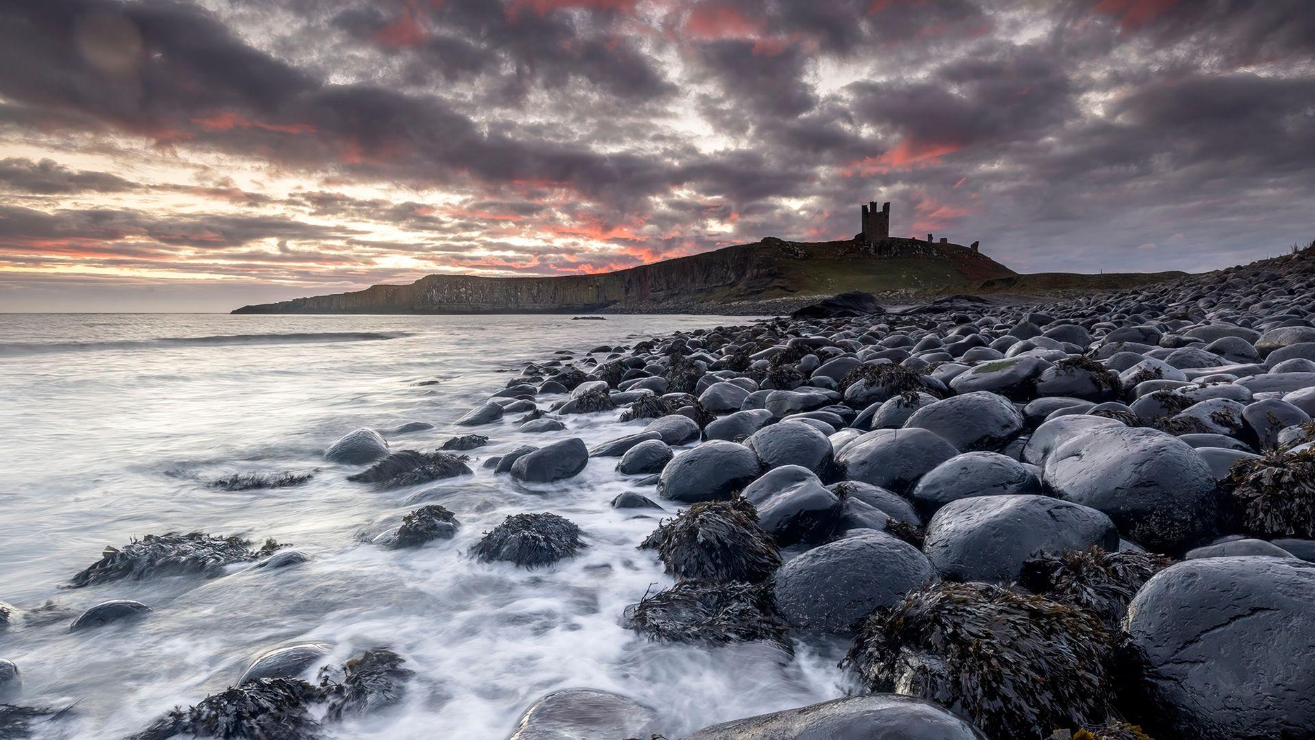 The sea washes up on large round black rocks, in the distance the ruins of a castle stand atop a cliff and the grey clouds are flecked with pink and orange streaks from a rising sun.