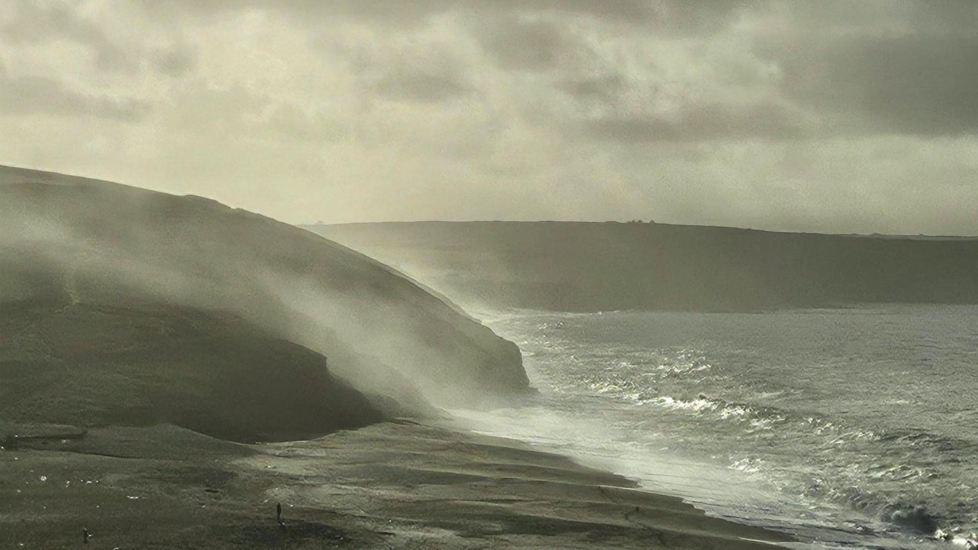Fine spray can be seen splashing up from the sea against rocks on the coast.
The land and sea appear grey with some glints of sun shining through.