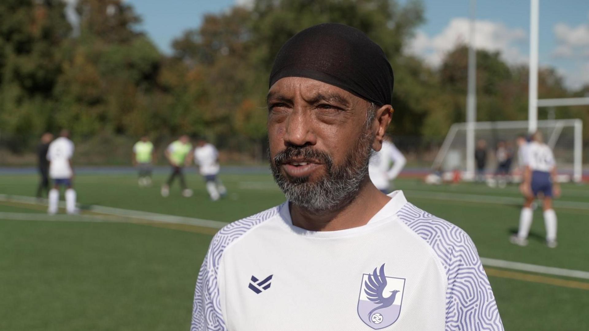 Jaspal is on the pitch wearing a white top. He has a a beard and black head covering on. Players can be seen in the background behind him.
