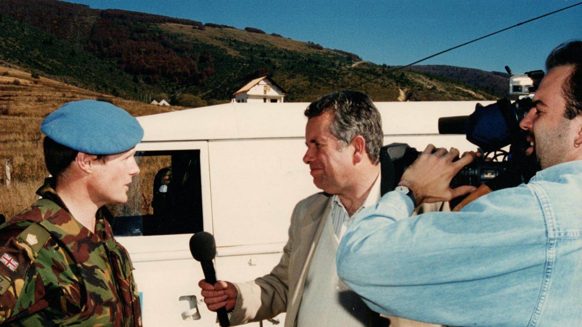 A young soldier in uniform being interviewed by a journalist with a microphone while being filmed by a cameraman in a blue shirt. They are standing next to a white van.