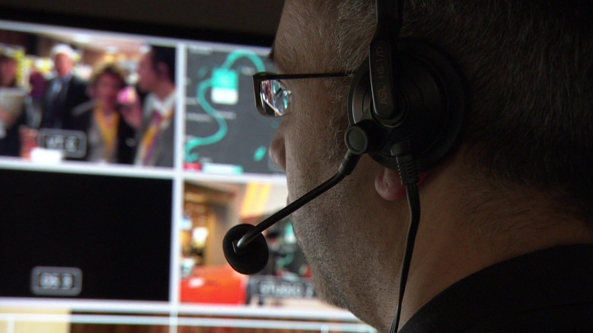 Close up shot of a man talking into a microphone with a backdrop of screens showing he is producing a TV programme 