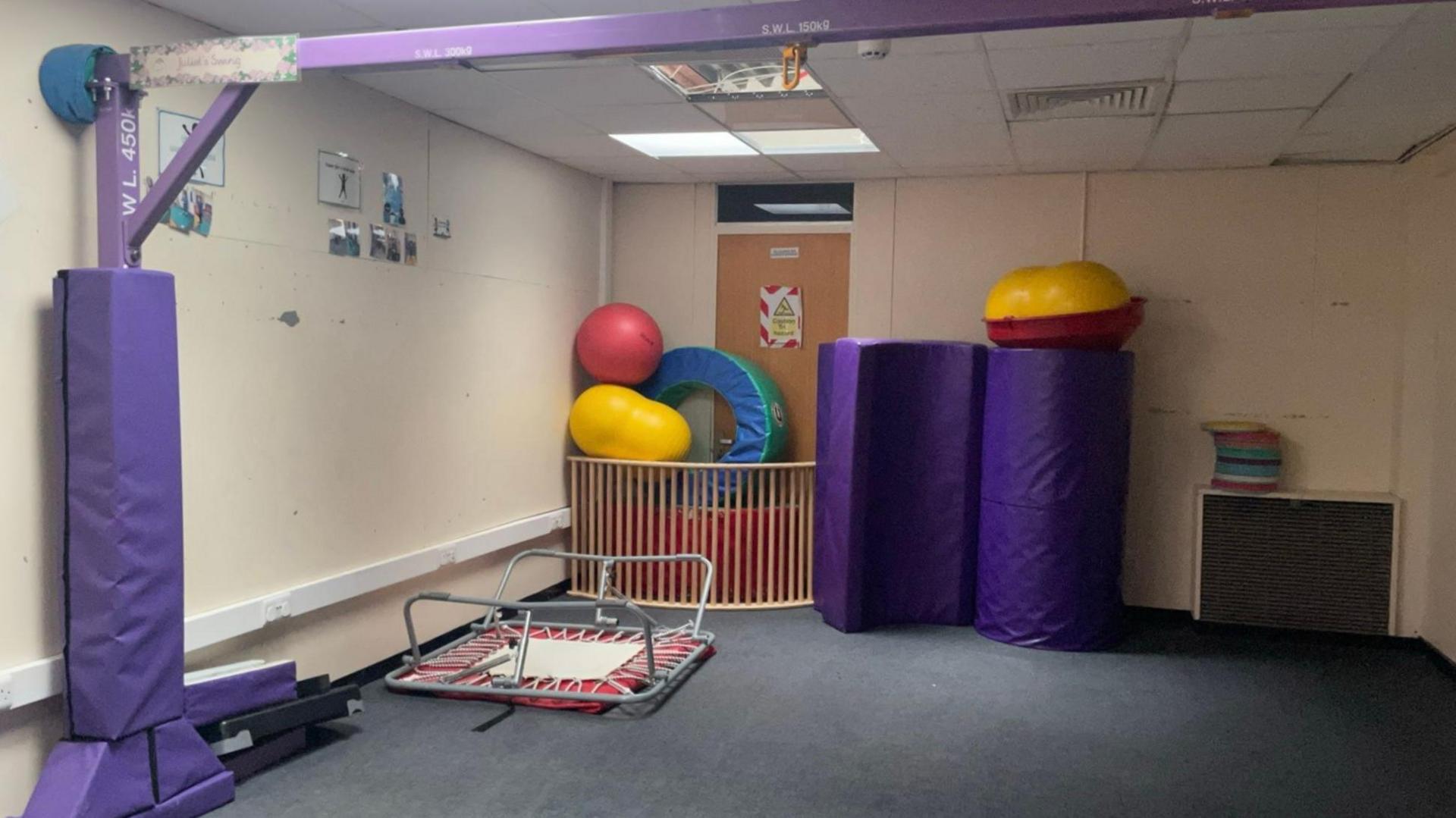 A dark, artificially lit room. A small trampoline is turned upside down. Colourful soft-play shapes and apparatus are stacked in a corner. Ceiling tiles look damaged and signs are stuck to the walls.
