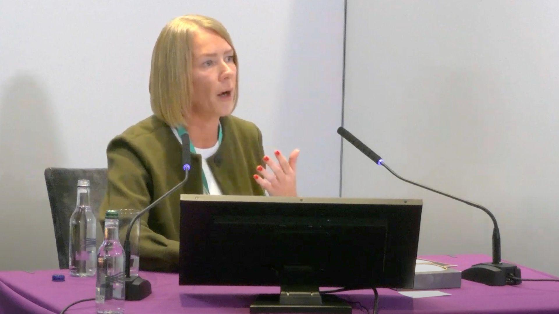 Laura Cozens gives evidence to the Lampard Inquiry. She sits at a purple table with two microphones and a computer monitor in front of her. She has shoulder-length blonde hair and wears a green jacket over a white t-shirt.