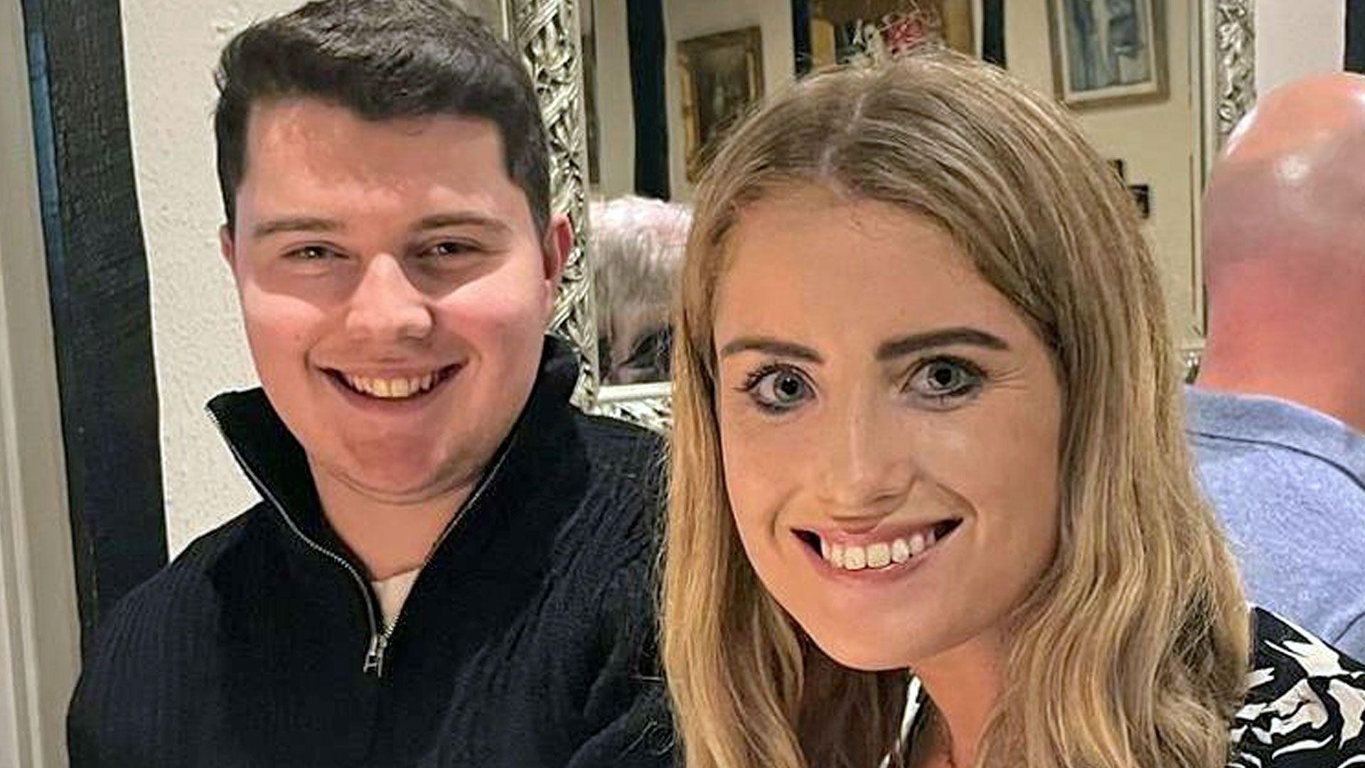 A young man and woman sit in a pub or restaurant, smiling at the camera. He wears a high-necked zip-up fleece, while the woman wears a top with a black and white pattern reminiscent of an animal print.