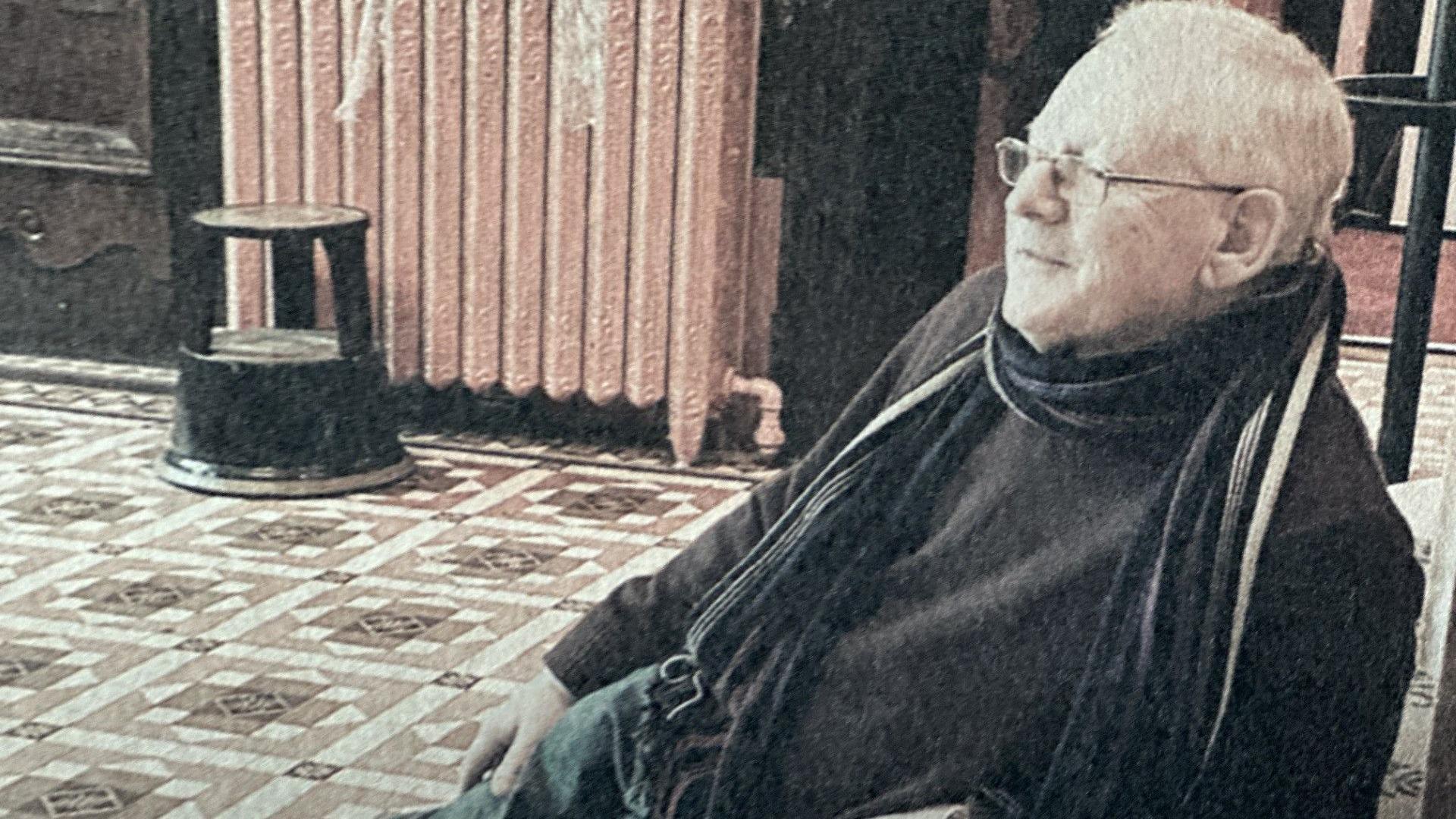 Mervyn Stokes, who has blond hair and wears glasses, sitting on a chair int eh foyer of the theatre. He's wearing a dark jumper and stripped scarf and the floor behind him is covered in patterned tiles.