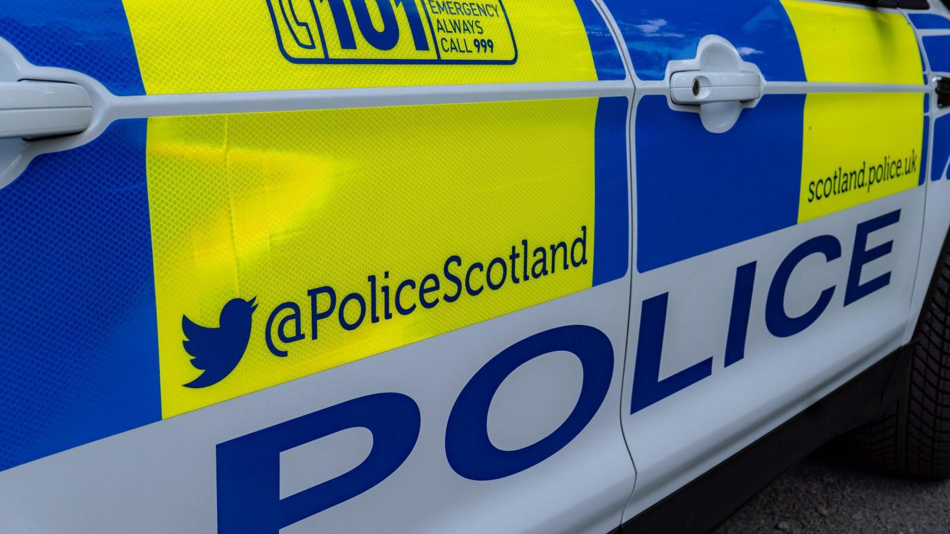 A close up of the exterior of a Police Scotland car. There are blocks of bright blue, yellow and white colours. The word "police" is in blue letters on a white background along the side of the car, and information on how to contact Police Scotland.