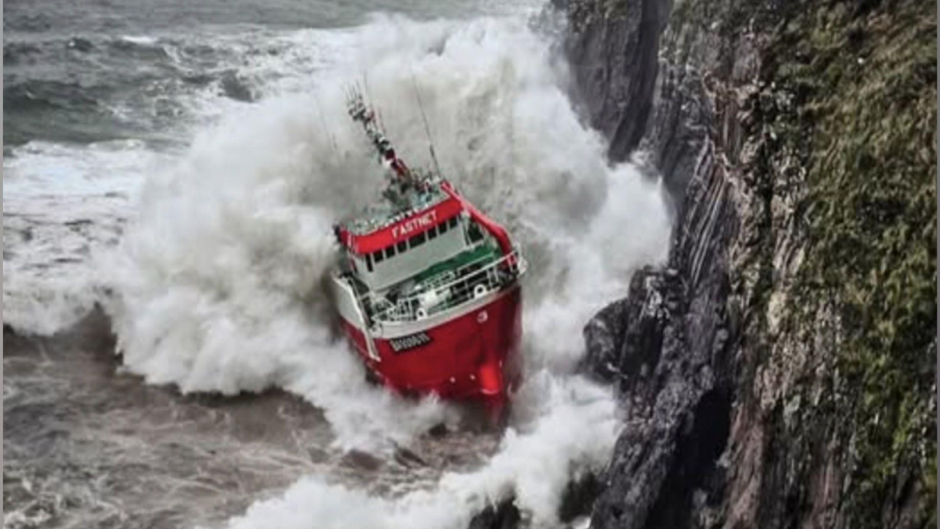 The image shows a red fishing vessel named 'Fastnet' appearing to hit a cliff edge during stormy weather. 