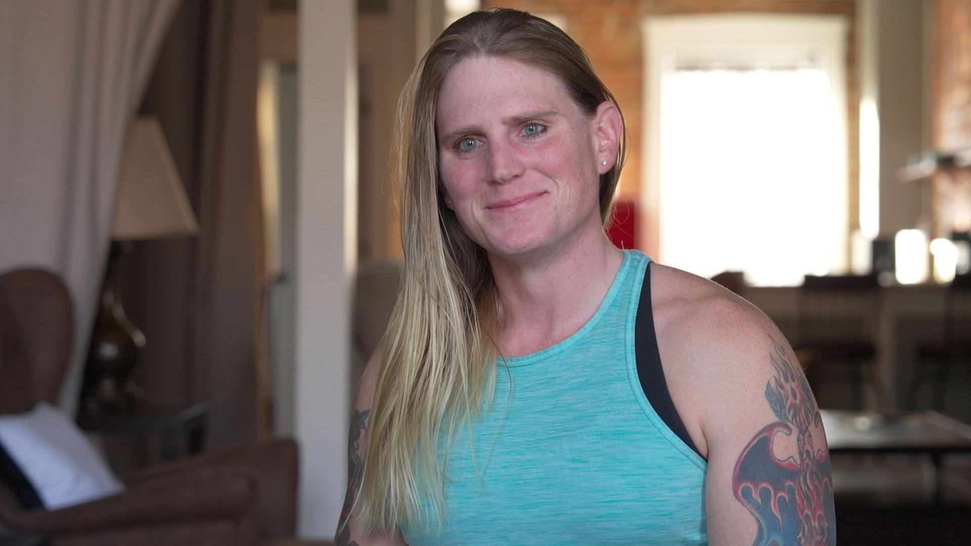 Kara's long straight blonde hair falls over her right shoulder. She is wearing a light blue vest top and has a tattoo of a phoenix rising, engulfed in flames, on her left arm.