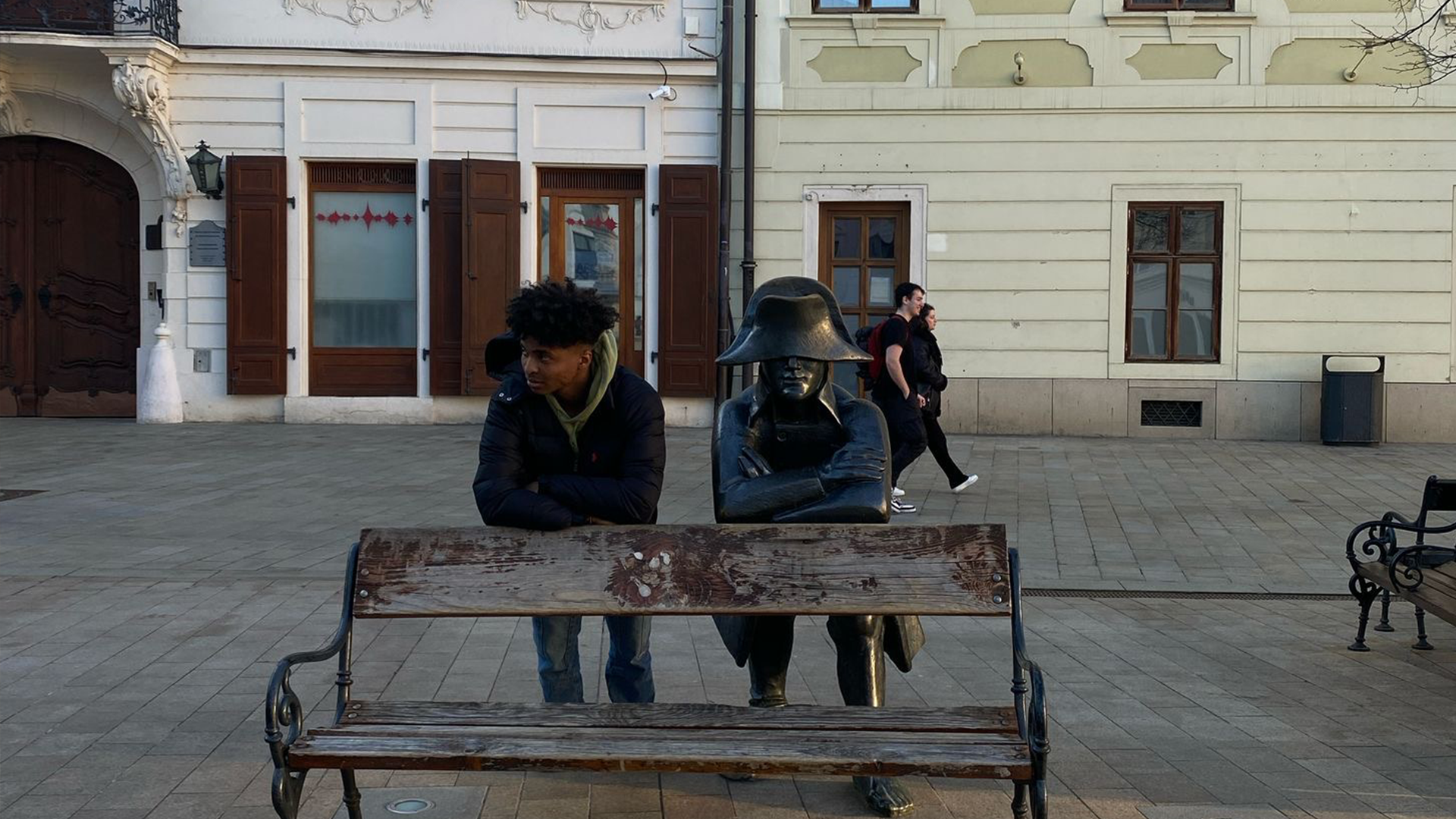 Luka Chijiutomi-Ghosh stands behind a bench next to a statue, in Bratislava, they are both holding a pose, leaning on the back of the bench with their arms crossed.