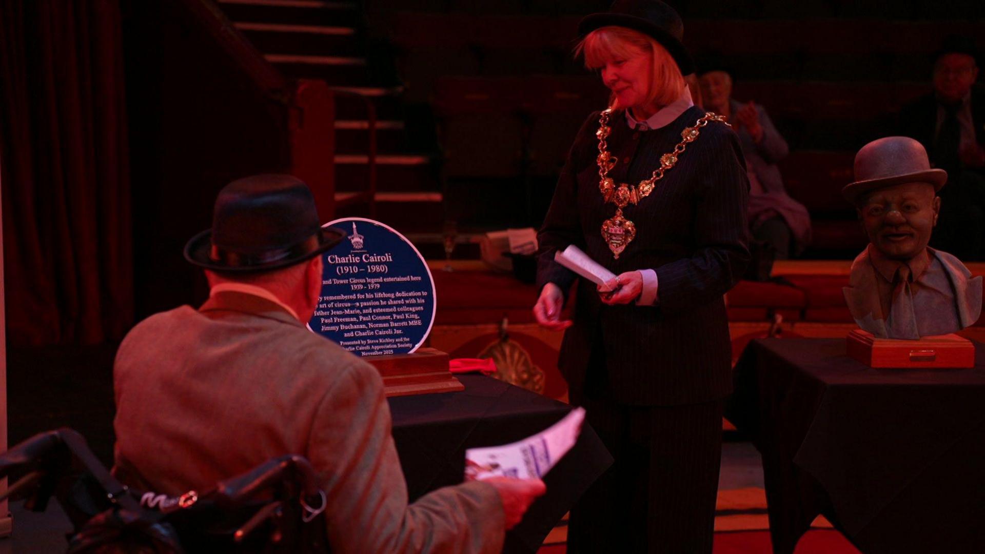 Blackpool's mayoress, wearing a bowler hat and heavy gold-coloured chain, joins Charlie Cairoli Junior, who sits in a chair - as they look at the round blue plaque for his father on a table. On the right is a bust of Charlie Cairoli wearing a bowler hat and suit.