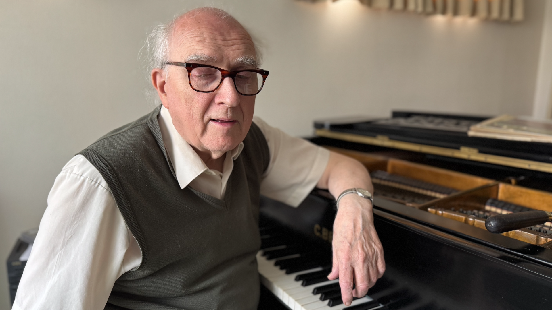 Paul Chrimes sitting at a grand piano wit his arm leaning on the top of it. He is balding with white hair and wearing glasses, a watch, a tank top and a short-sleeved white shirt.