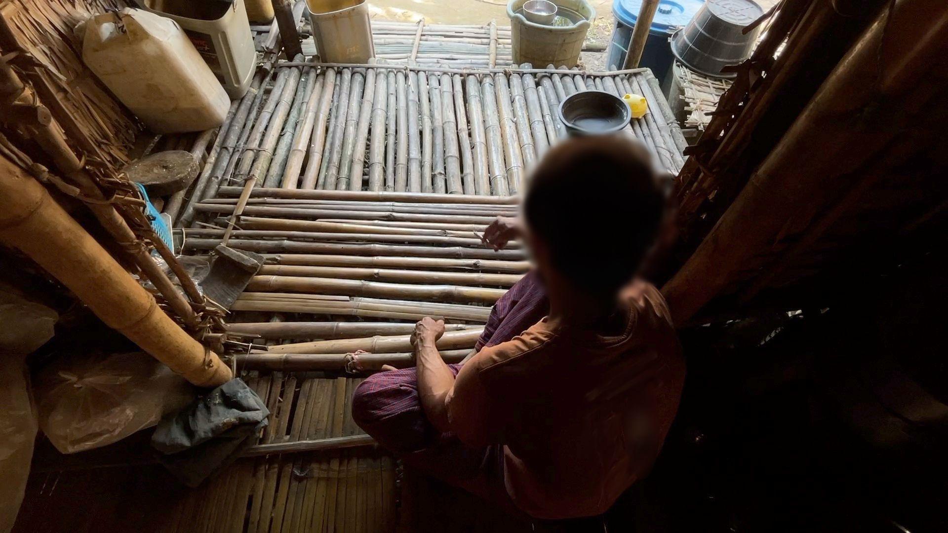 Image showing Zeya with his back to the camera, sitting cross legged on a floor made of bamboo poles, holding a cigarette in his right hand. His head is blurred and there are several water containers in buckets in the background. 