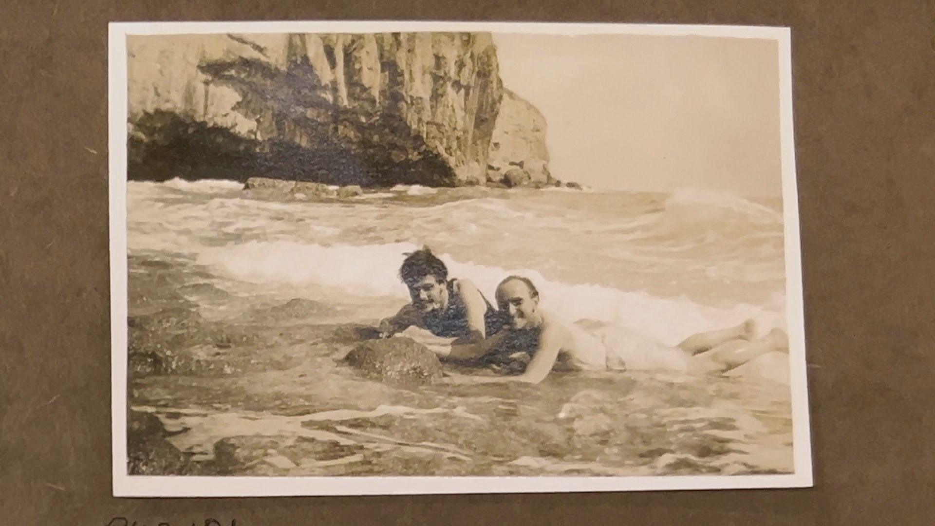 A black and white photograph of David Brynley and Norman Notley laying on their front in the shallows of the sea in swimming trunks. Behind them is a large cliff.