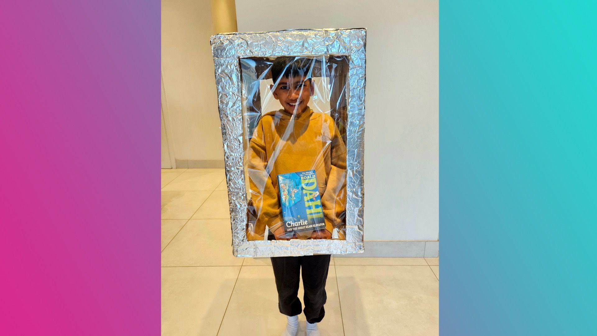 A boy wearing a yellow hoodie and black trousers stood in a box made out of see-through plastic with foil edges. He's holding a copy of Charlie and the Great Glass Elevator by Roald Dahl inside the box.