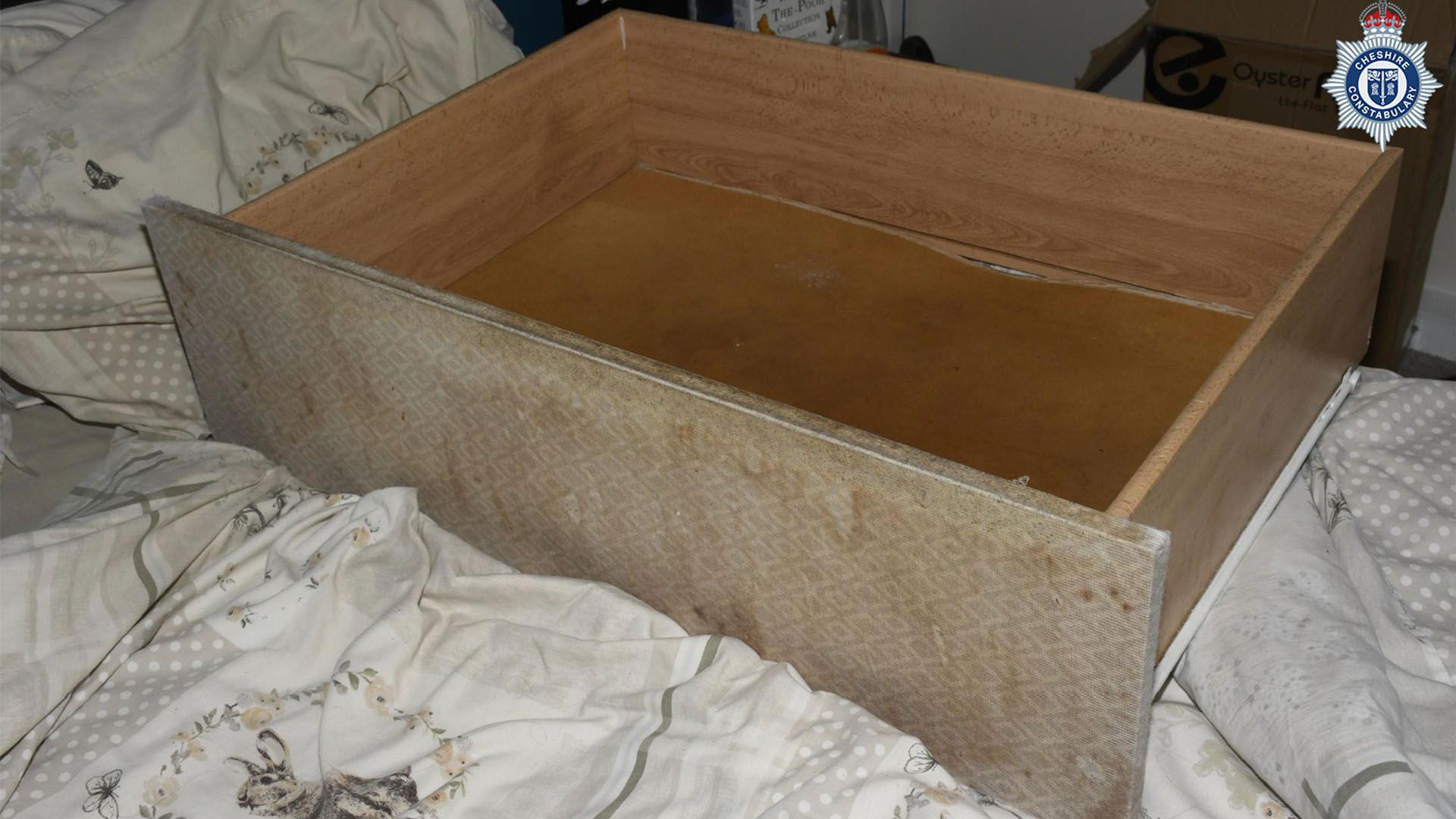 A wooden draw from the divan bed where the child was kept, pulled out and placed on top of a bed. The cover of the drawer is dated and covered in brown marks. 