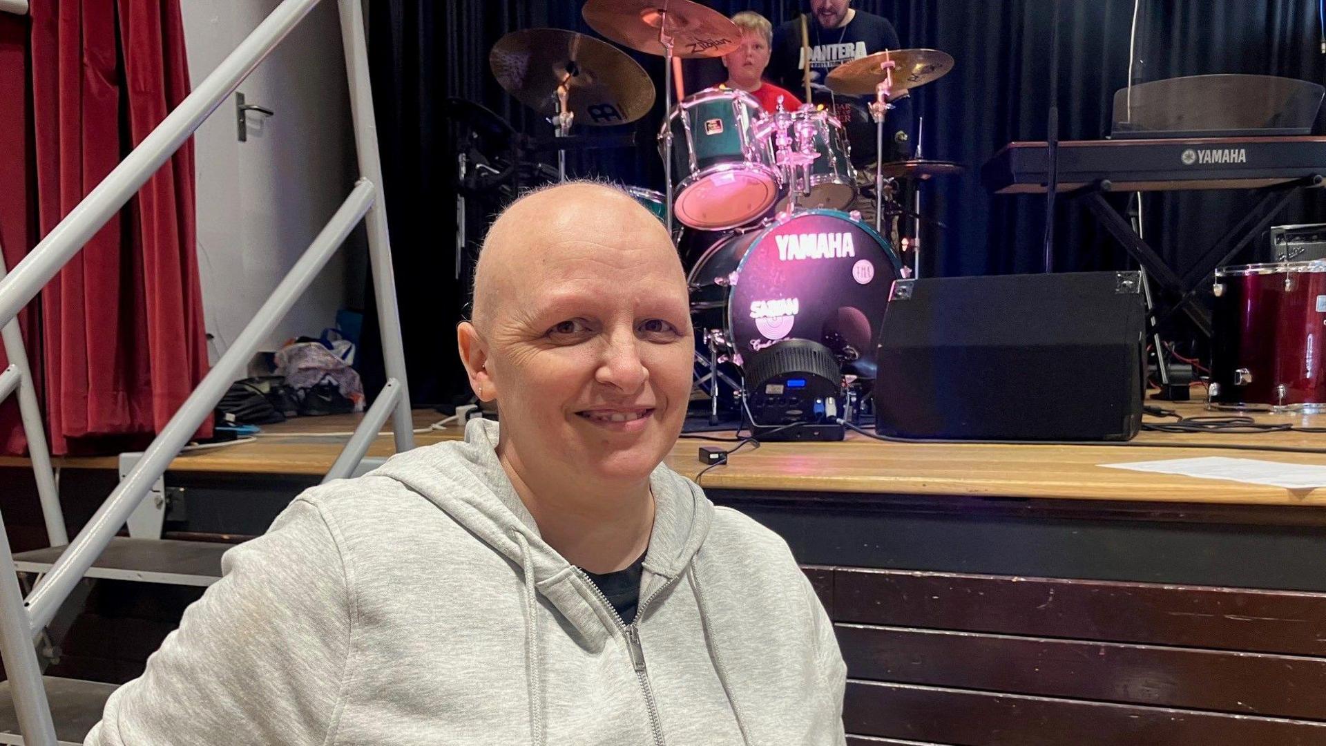 Dawn standing in front of her son sitting at a drum kit, she's lost her hair due to chemotherapy. She is at the bottom of steps leading up to the stage Jack's drumkit is on. He is wearing a red T-shirt and a man is visible in a black T-shirt with white writing standing slightly behind him. 