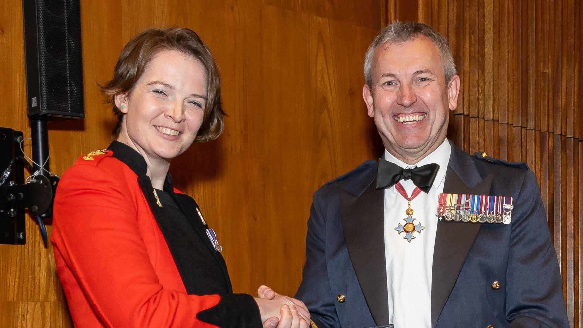 A woman smiles as she shakes hands with a man in a suit who has medals on his chest.