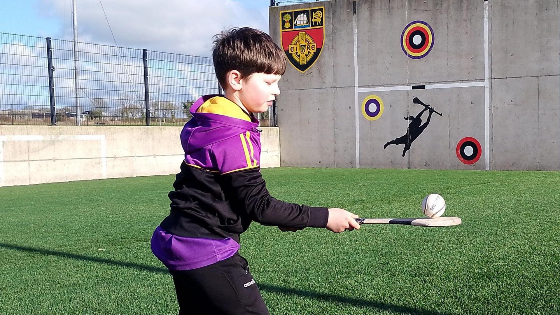 Fionn O'Neill playing hurling.  The eight-year-old is balancing a sliotar on a hurling stick in a practice pitch.   He has short dark hair and is wearing the black, purple and yellow sportswear of Carryduff GAC