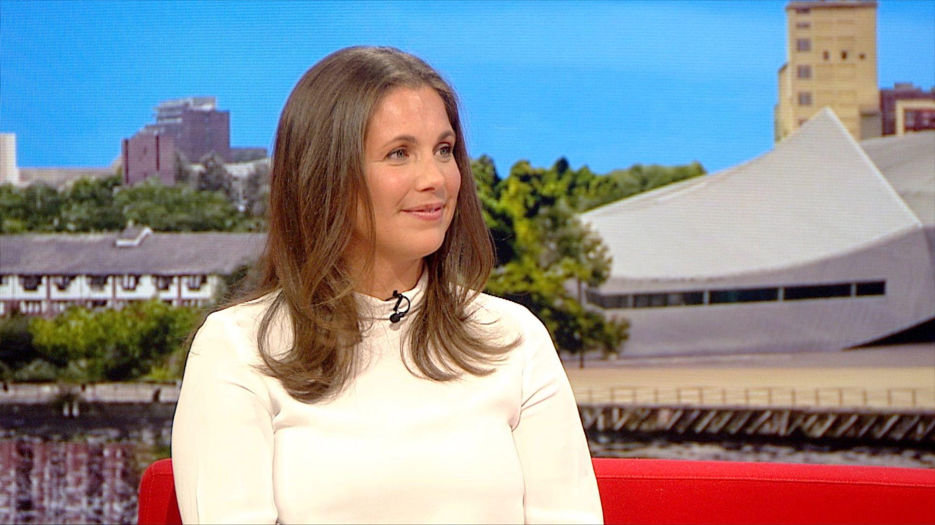 A mid-shot of Lindsey Burrow with long brown wavy hair and a white polo neck