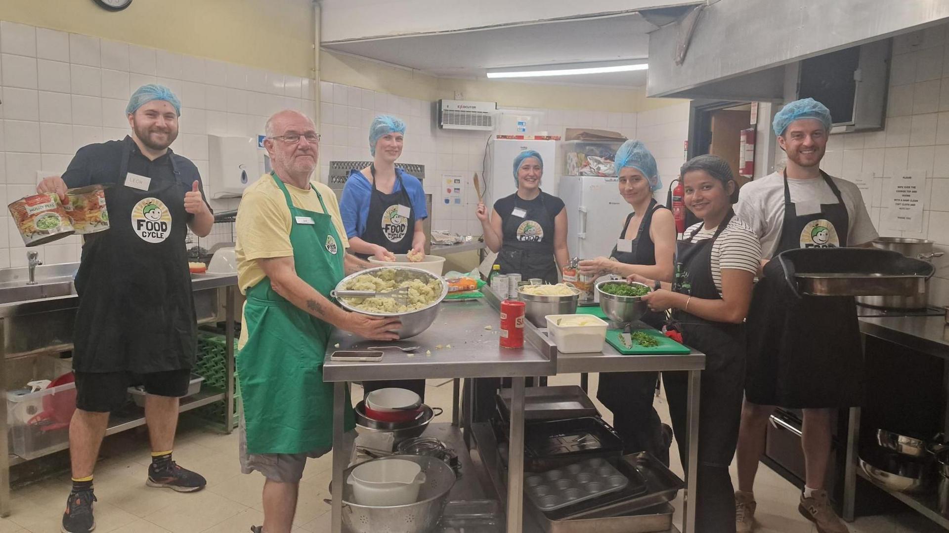 Volunteers wearing aprons and hairnets prepare vegetables and ingredients around a central worktable in a community kitchen. Ian White is stood with them with glasses, a yellow t-shirt and green apron.
