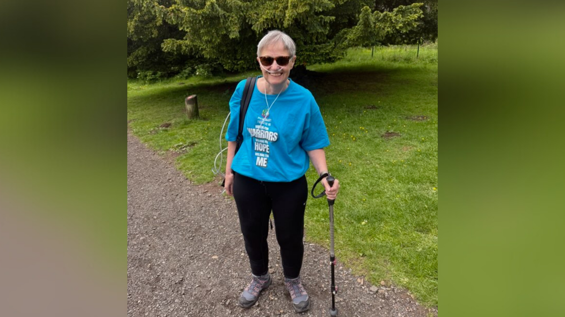 Una McMillan standing in a blue t-shirt and black trousers holding a hiking pole. She has oxygen tubes and carries an oxygen tank. She is in a park.