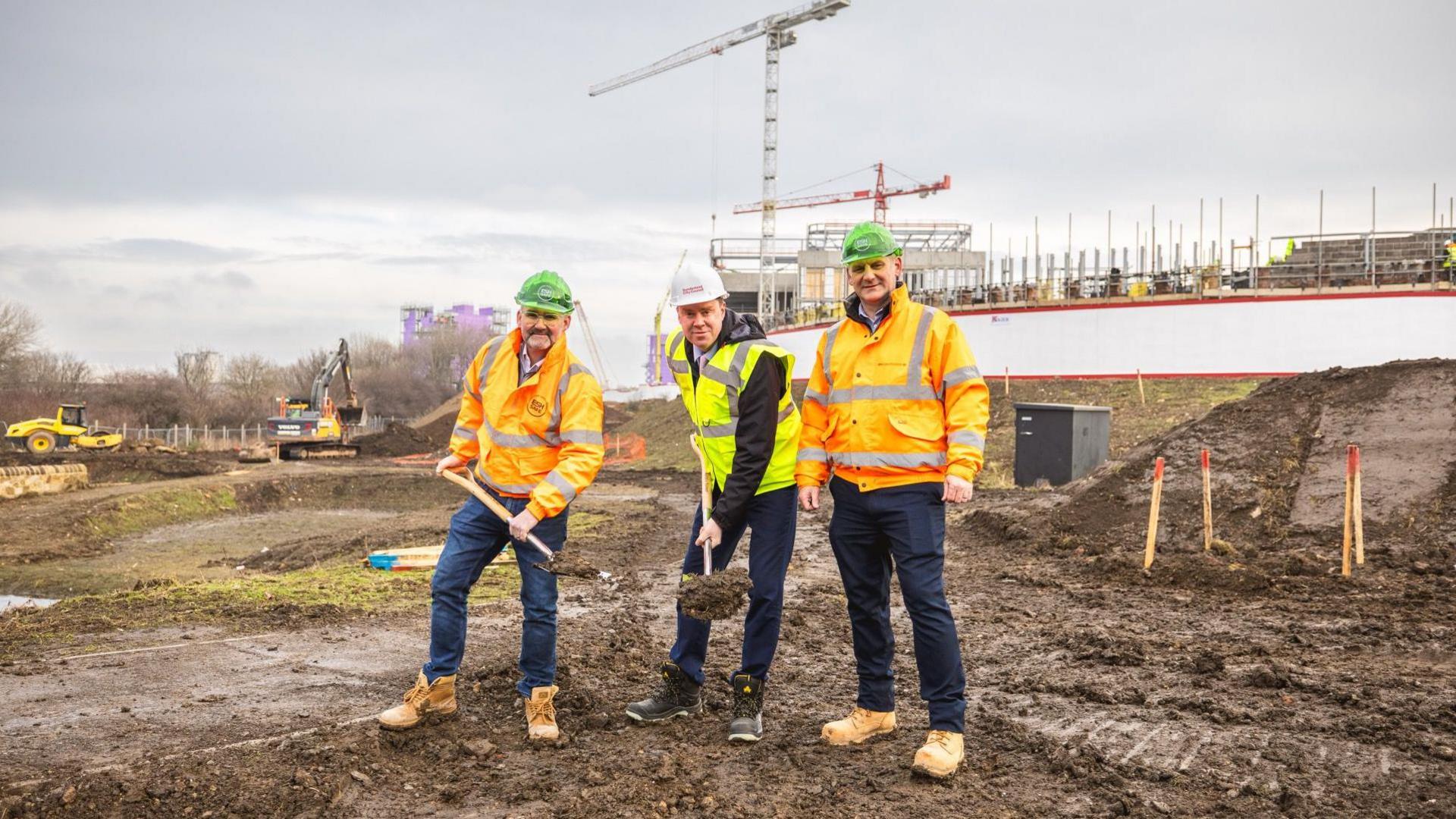 (L-R) Andy Radcliffe, chief executive of Esh Construction, councillor Kevin Johnston and Stephen McClean, construction manager at Esh Construction at the building site of the new park. Mr Radcliffe and Johnston are both holding spades which have been used to dig up earth. Excavators can be seen in the background and the ground is muddy.