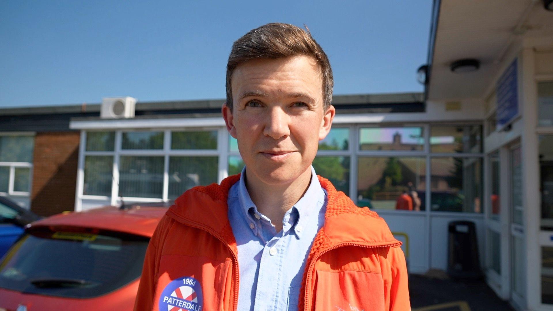Dan Jones looks at the camera. He has short brown hair and is clean shaven. He is wearing a blue shirt and orange mountain rescue jacket. Behind him is a single storey hospital building