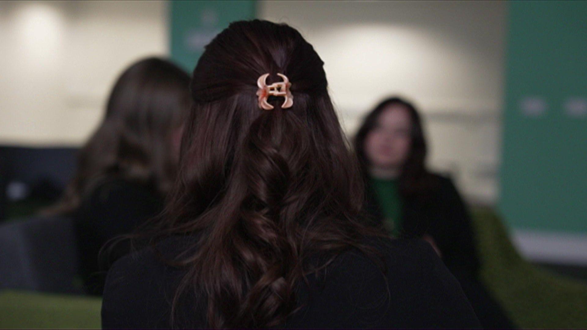 Three dark haired women are sitting talking to one another, the shot is out of focus. The back of one's head with a pink hair clip is in focus in the foreground. They are sitting in a green room.