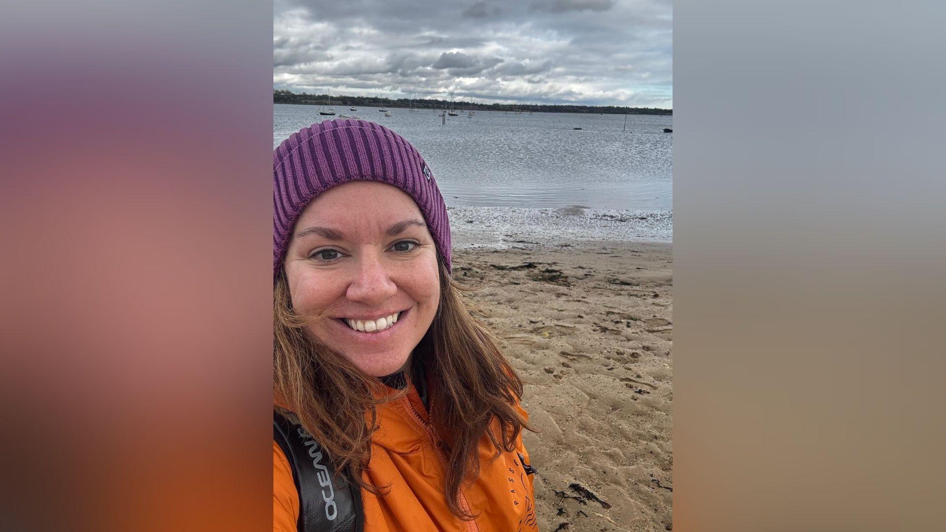 Mairead wears a cerise bobble hat and orange Dryrobe, and is standing on a beach. She is smiling.
