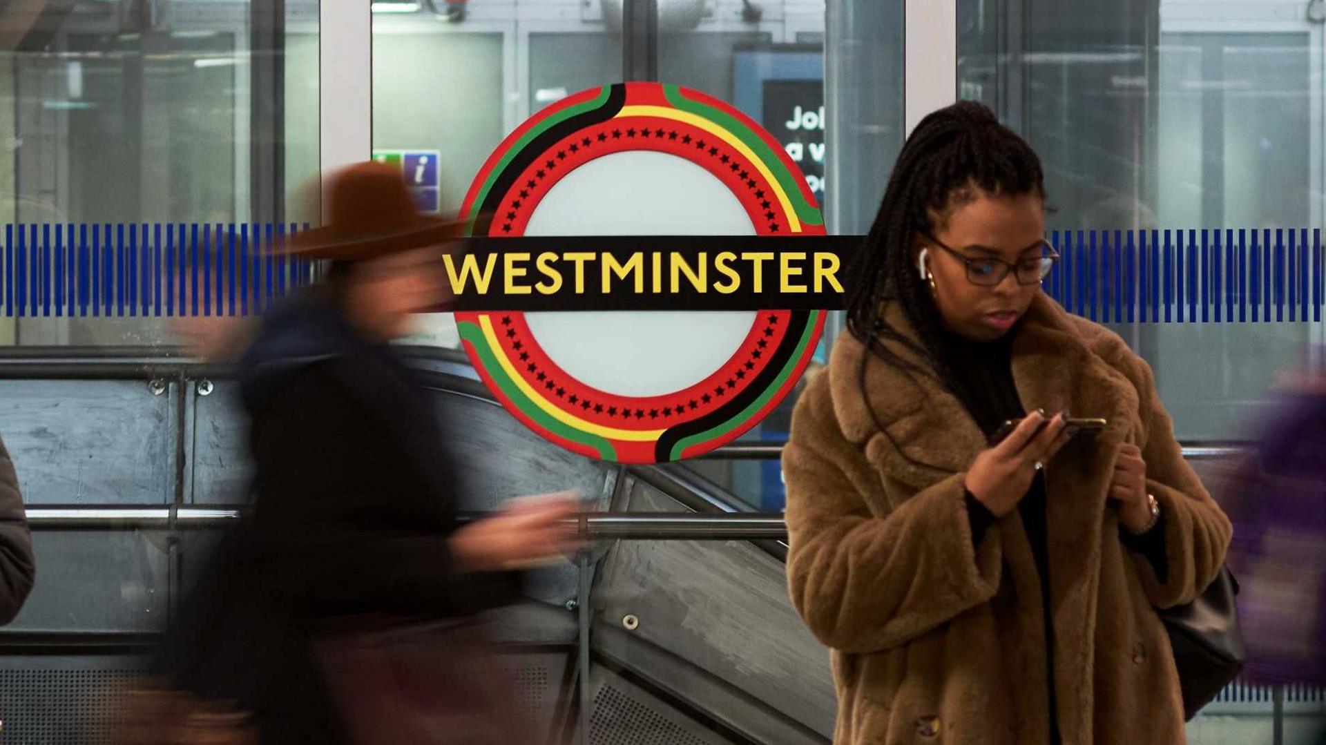 A Westminster tube station roundel in the Pan-African colours, with a lady in a faux fur coat standing in front of the roundel looking at her phone and another person out of focus walking along the platform. 