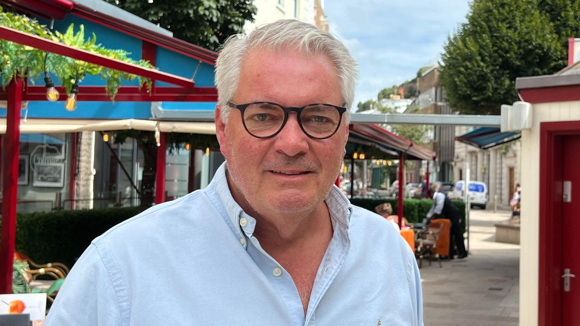 Bar owner Davis Voisin outside the Bar Levant al fresco bar and restaurant, wearing a light blue open collared shirt and round glasses. He has grey hair and light stubble.