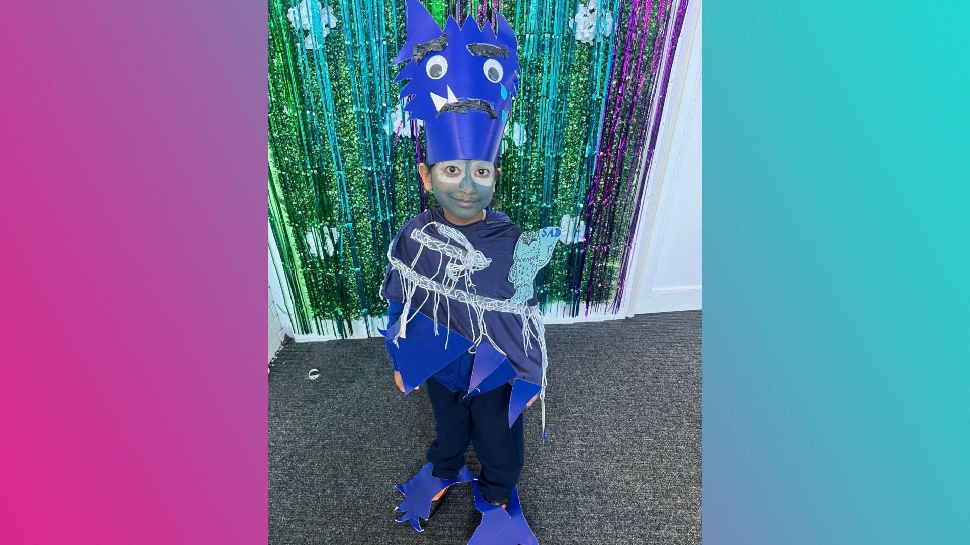 A child wearing a blue top, blue trousers and a blue headpiece which has the face of a monster on it. The monster has big eyes and two big teeth. The girl is standing in front of a green, blue and purple sparkly decoration on the door.