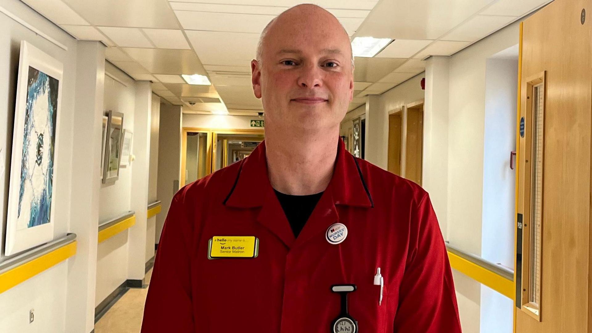 Mark Butler, a bald man, wearing a red matron uniform in a hospital corridor.