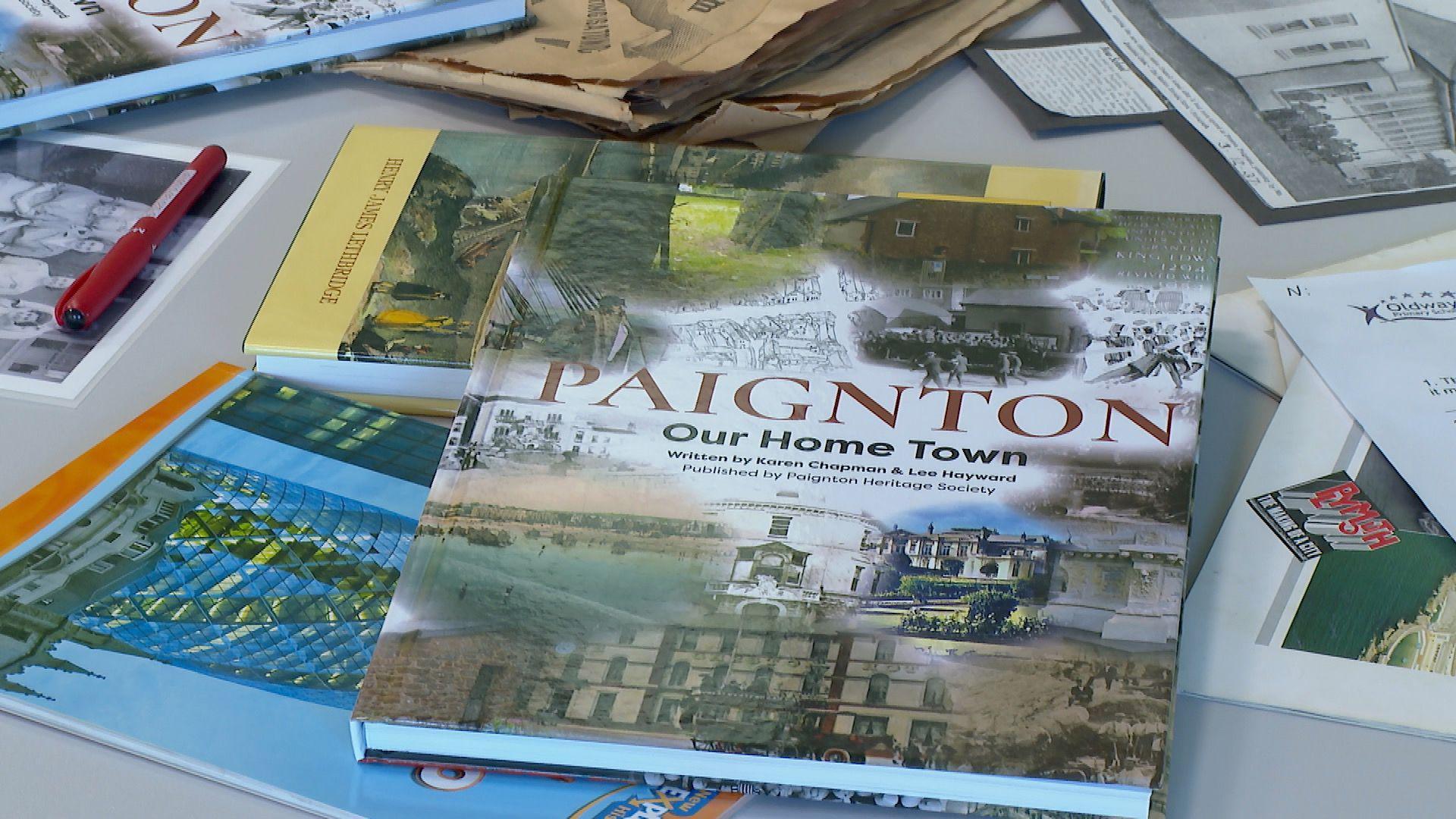 A book on a table, surrounded by other books and magazines. The book has its title 'Paignton - Our Home Town' on the front, along with photos of Paignton.