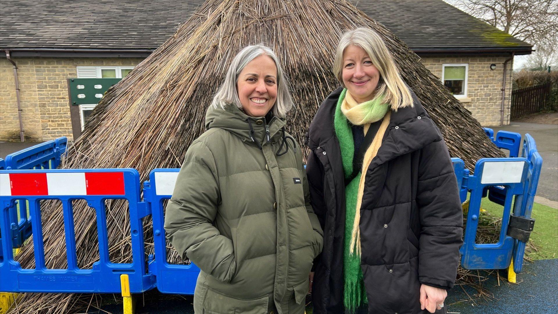 Two women standing in front of the thatched hut with blue fencing around it.