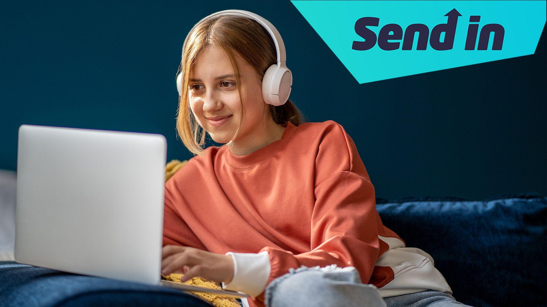 A girl sits on a sofa looking at her laptop and smiling. She is wearing a coral jumper, jeans and a pair of over ear headphones. There is a graphic in the top right corner that says "send in".