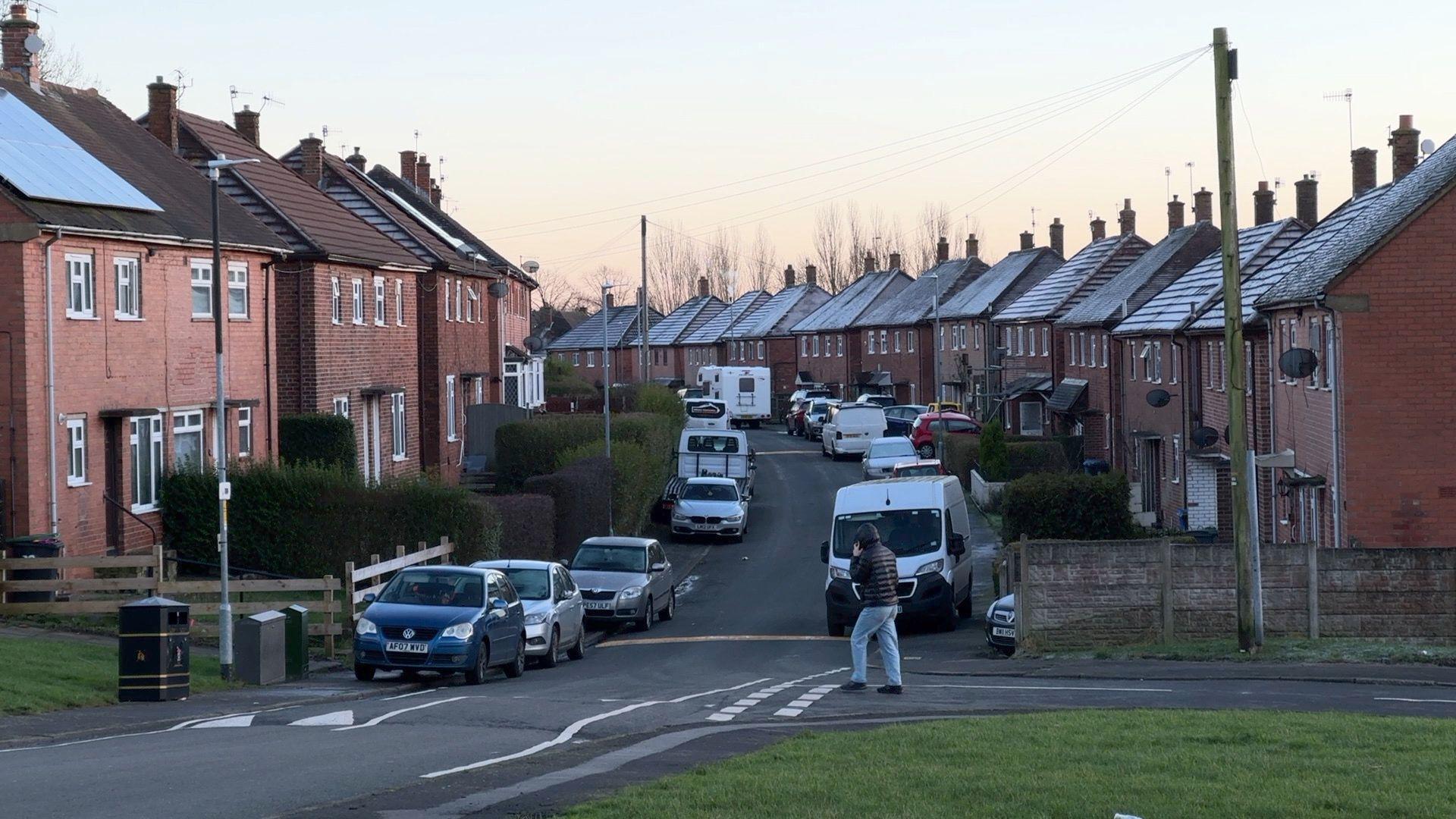 A residential street in Stoke-on-Trent, with houses on either side of the road which curves round to the left. There are cars and vans parked on both sides of the street and a speed bump can be seen with road signs. A person is crossing the road.