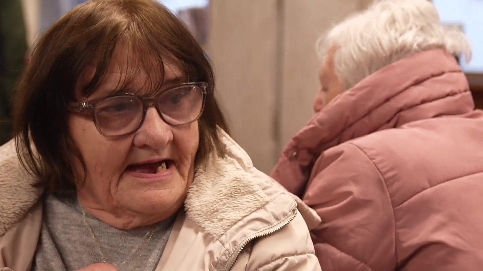 Carol in a warm hub in Tower Hamlets. She has brown hair and glasses. She wears a grey top and cream coat.