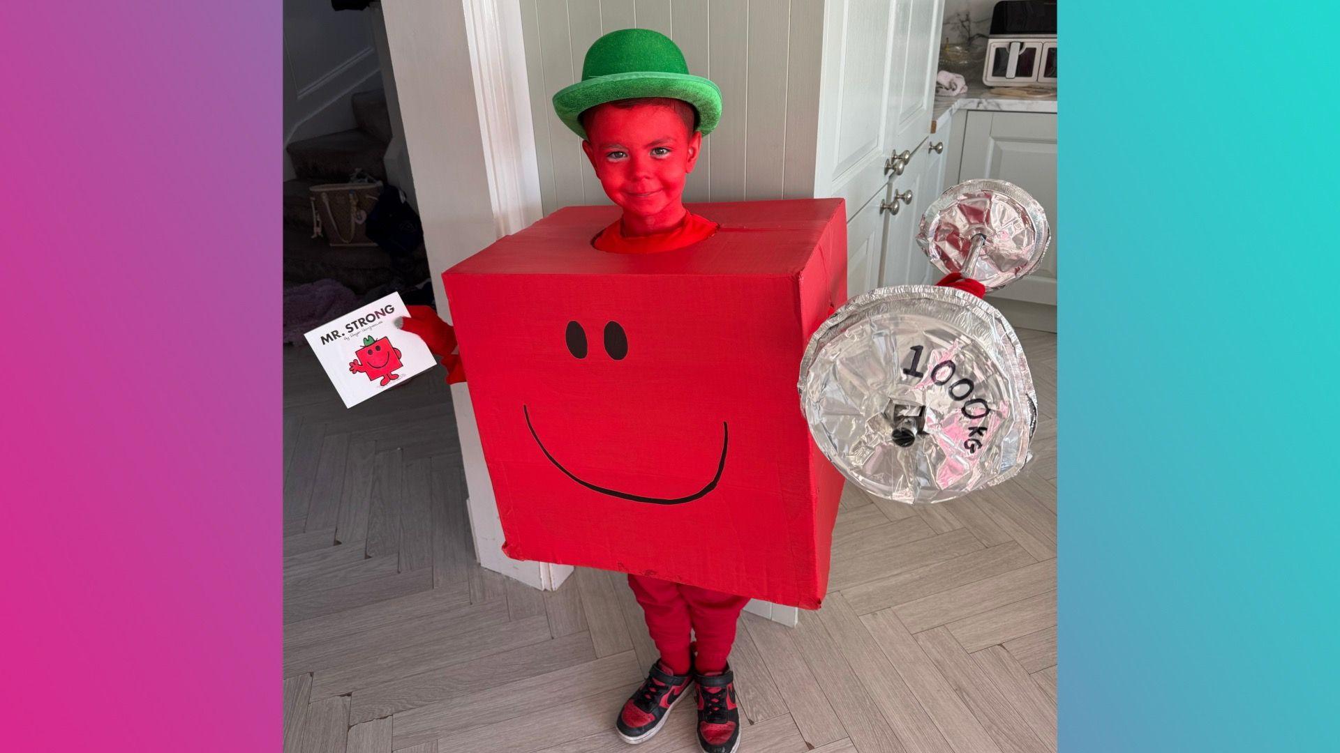 A boy wearing a red cube shaped costume with a smiley face on it and a green hat. He's wearing red face paint, red trousers and red shoes. He's carrying a pretend weight that says 1,000 kilograms on it in one hand, and the Mr Strong book in the other.