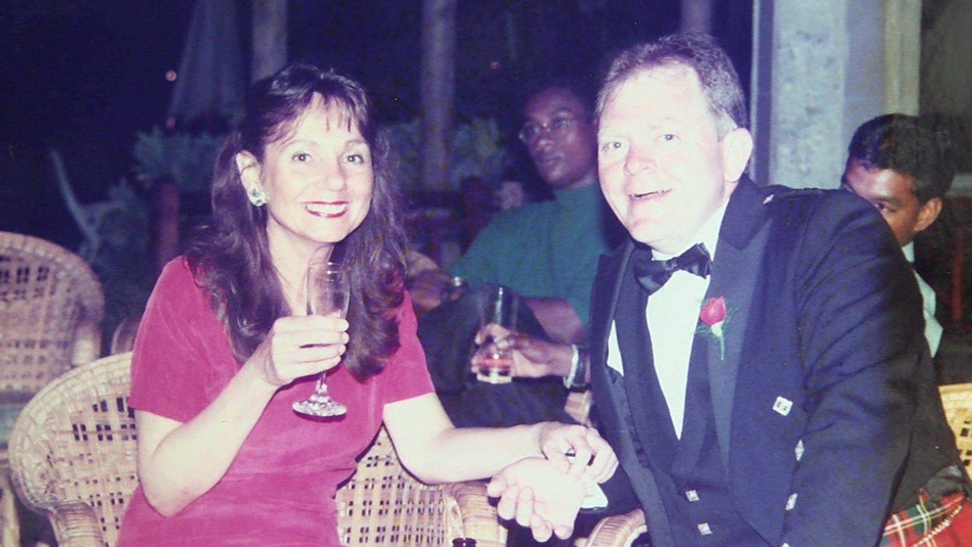 An old photograph of Dave and Carole when they were younger. The couple are dressed smartly with Dave wearing a black tie and Carole a red dress. They are holding hands and sat down while Carole is also holding a glass of champagne.