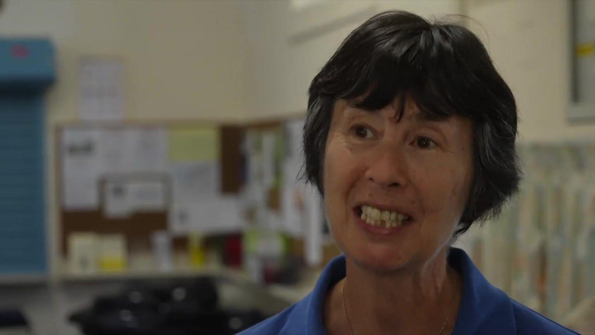 A woman wearing a blue polo shirt. She is stood looking at a camera and smiling. In the background are noticeboards covered with paper.